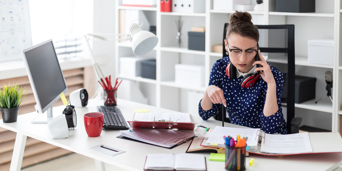 A young girl in glasses sits in the office at a computer desk and is talking on the phone. Before her on the table lie folders with documents.