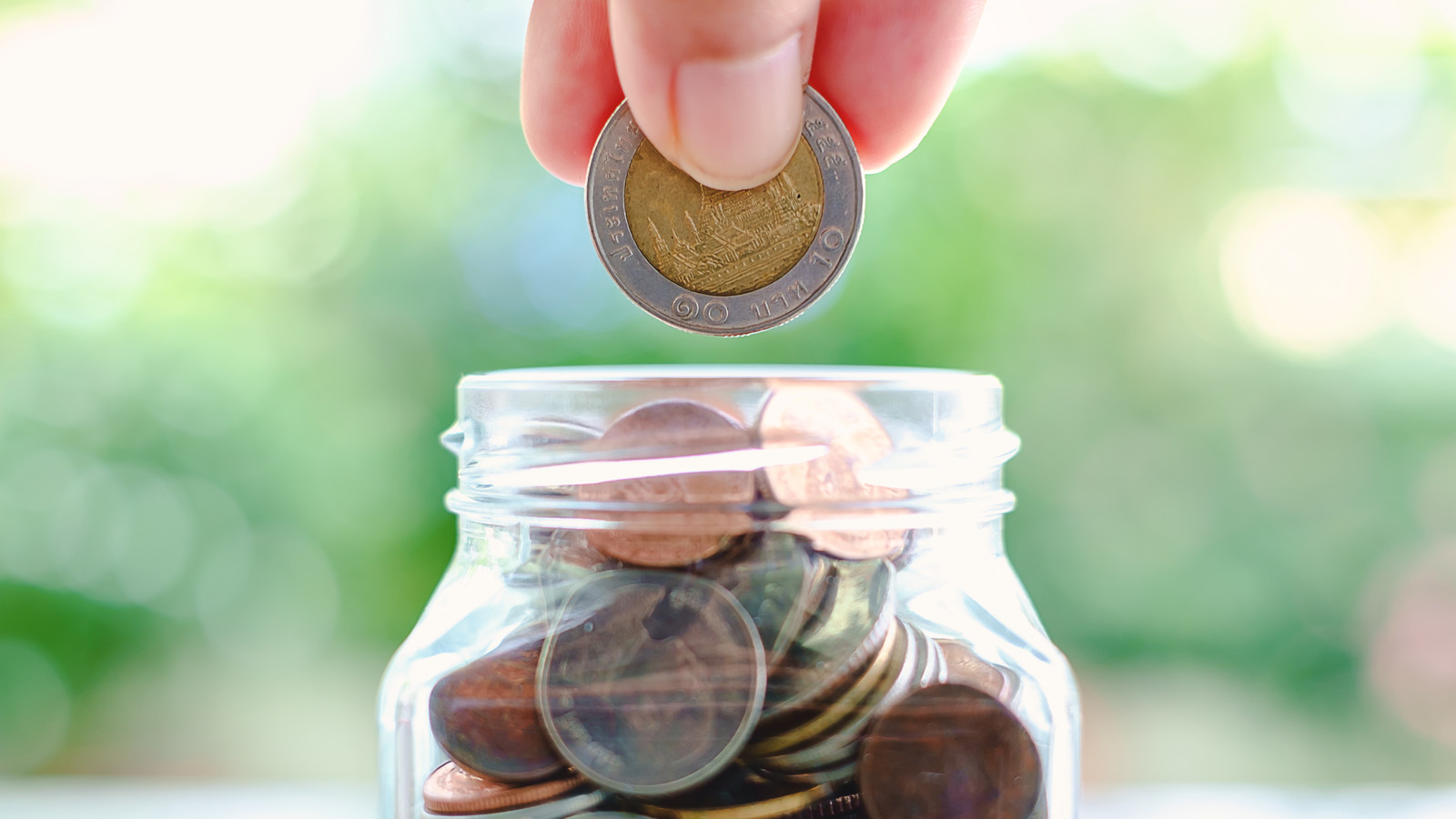 Hand saving a coin into the glass jar on blurred green natural background for business and finance concept